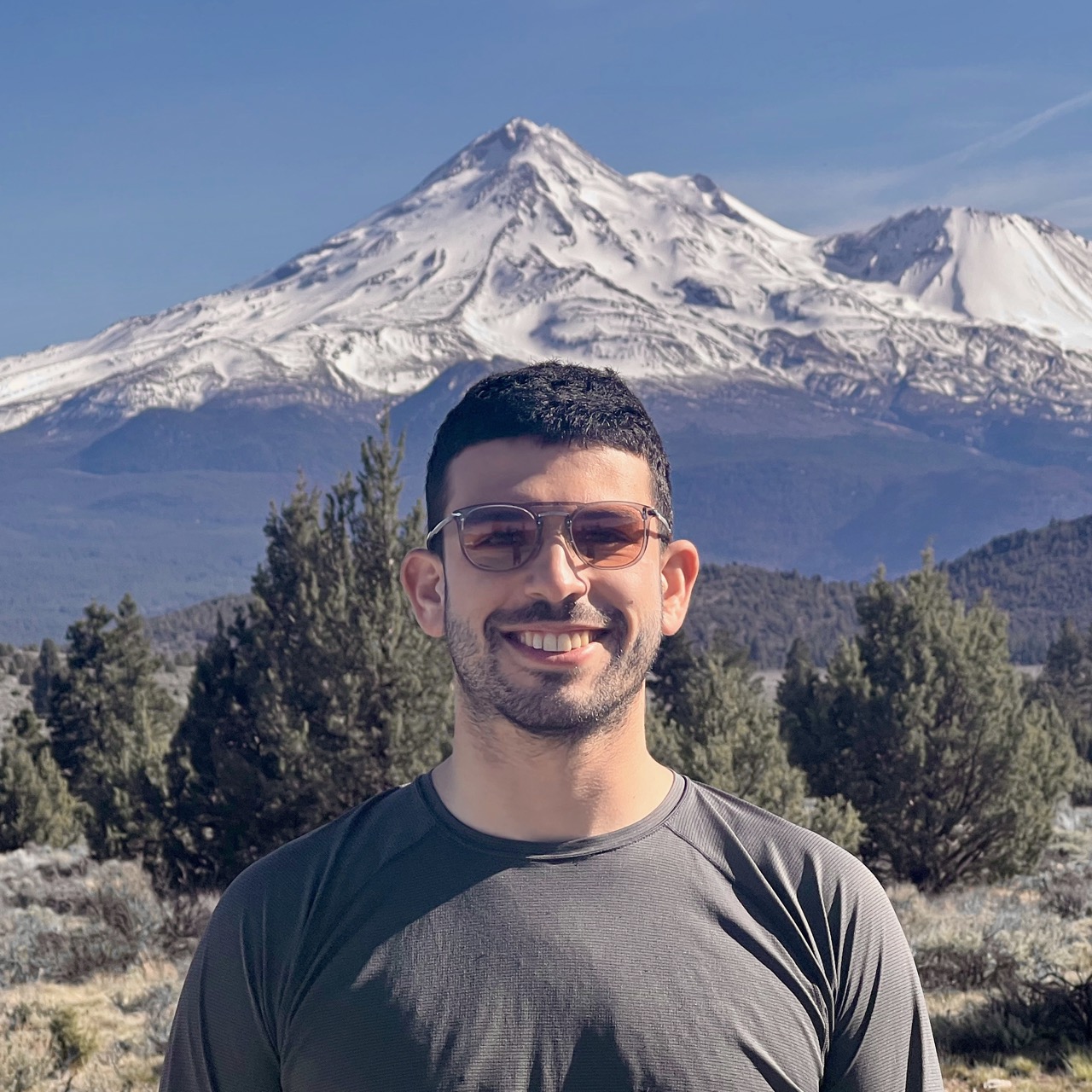 Portrait of me, smiling, wearing sunglasses and a gray shirt. There's some scrubland in the midground. The North side of Mount Shasta rises behind me.