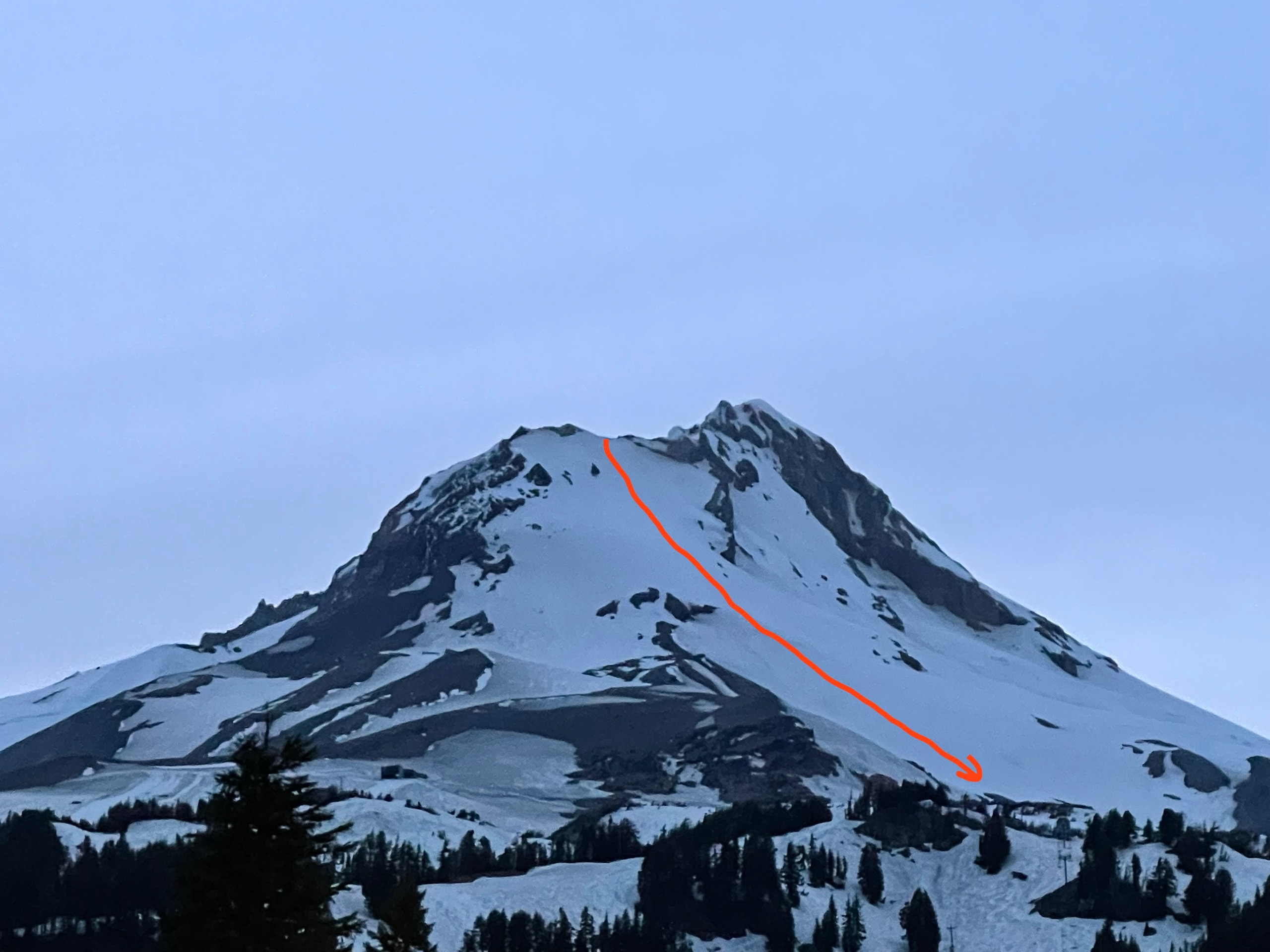 A photo of Wy'east from the southeast at dusk. The upper mountain is blanketed in snow, and the snow fields break up and become patchy at lower elevations, revealing brown volcanic rock. A red line outlines our planned route through the snow.